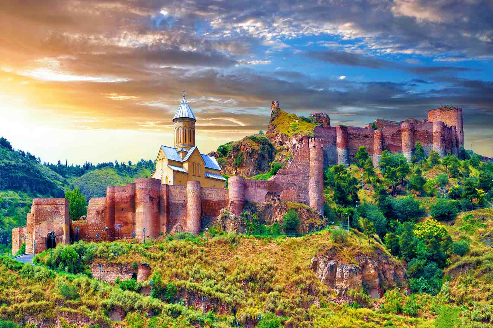 Metekhi Church and fortress ruins on hillside in Georgia at sunset with dramatic sky and green vegetation